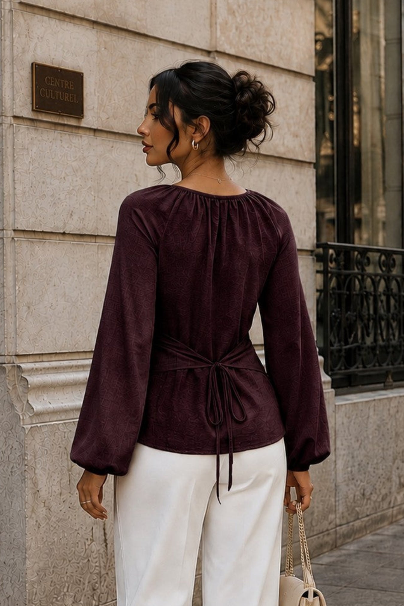 A woman stands on a city sidewalk wearing a deep burgundy long-sleeve top with a drawstring waist and white wide-leg pants, holding a small beige handbag.
