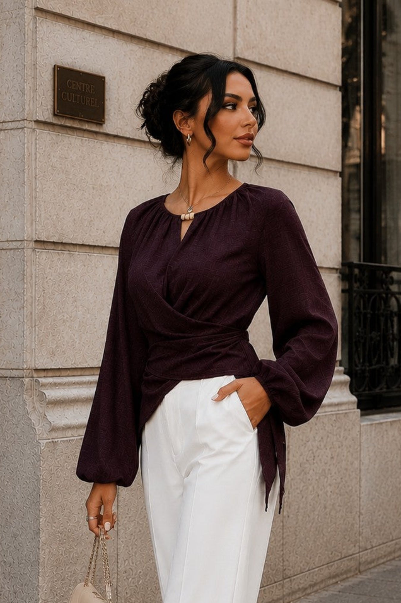 A woman stands on a city sidewalk wearing a dark plum wrap-style long-sleeve blouse and white wide-leg pants, holding a small beige handbag. The background shows a light stone building facade with dark windows.