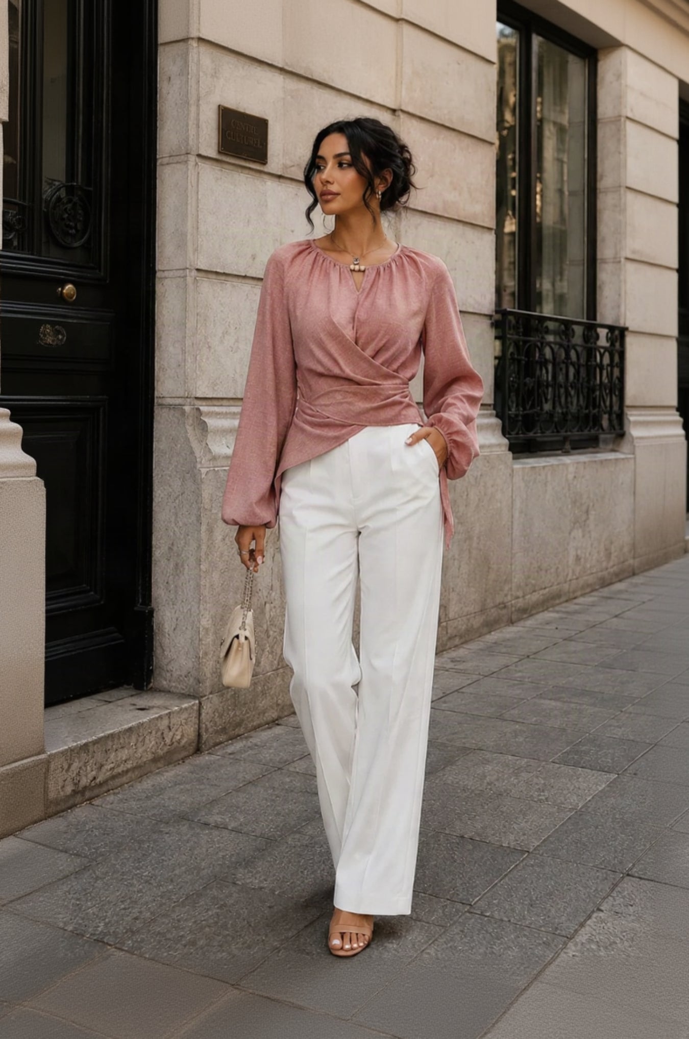 A woman stands on a city sidewalk wearing a dusty rose long-sleeve wrap-style blouse with white wide-leg trousers, holding a small beige handbag.