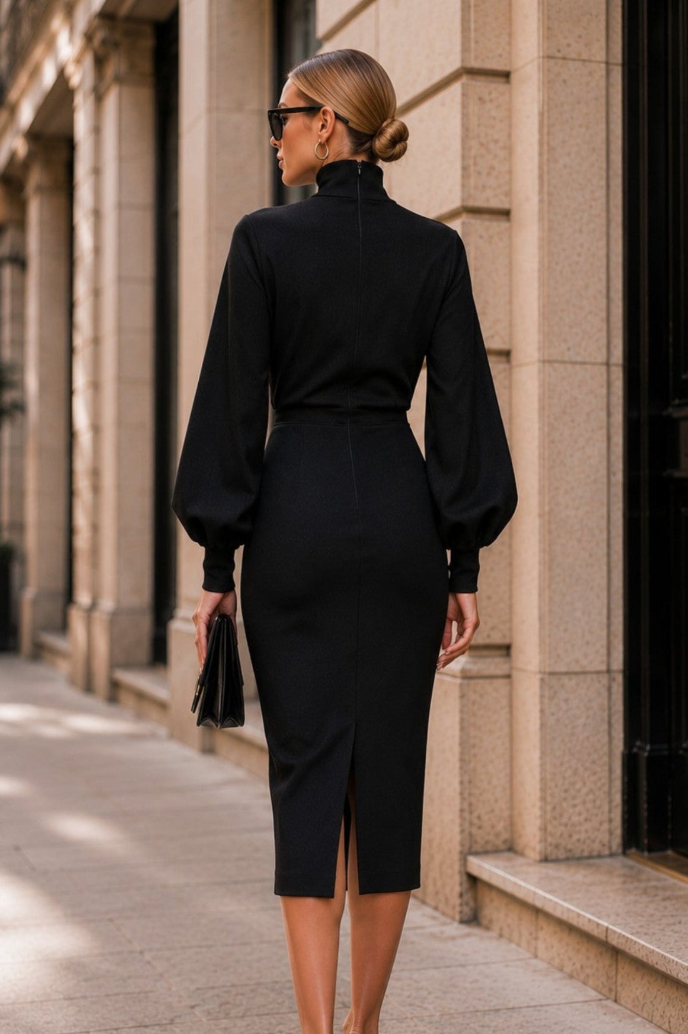 A woman wearing a black long-sleeve mock-neck pencil dress with balloon sleeves and a front slit, walking on a city sidewalk.