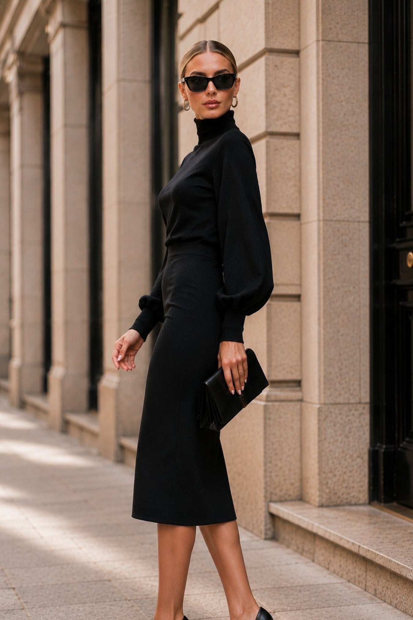 A woman stands on a city sidewalk wearing a black long-sleeve midi dress with balloon sleeves, black pointed-toe heels, and holding a black clutch bag.