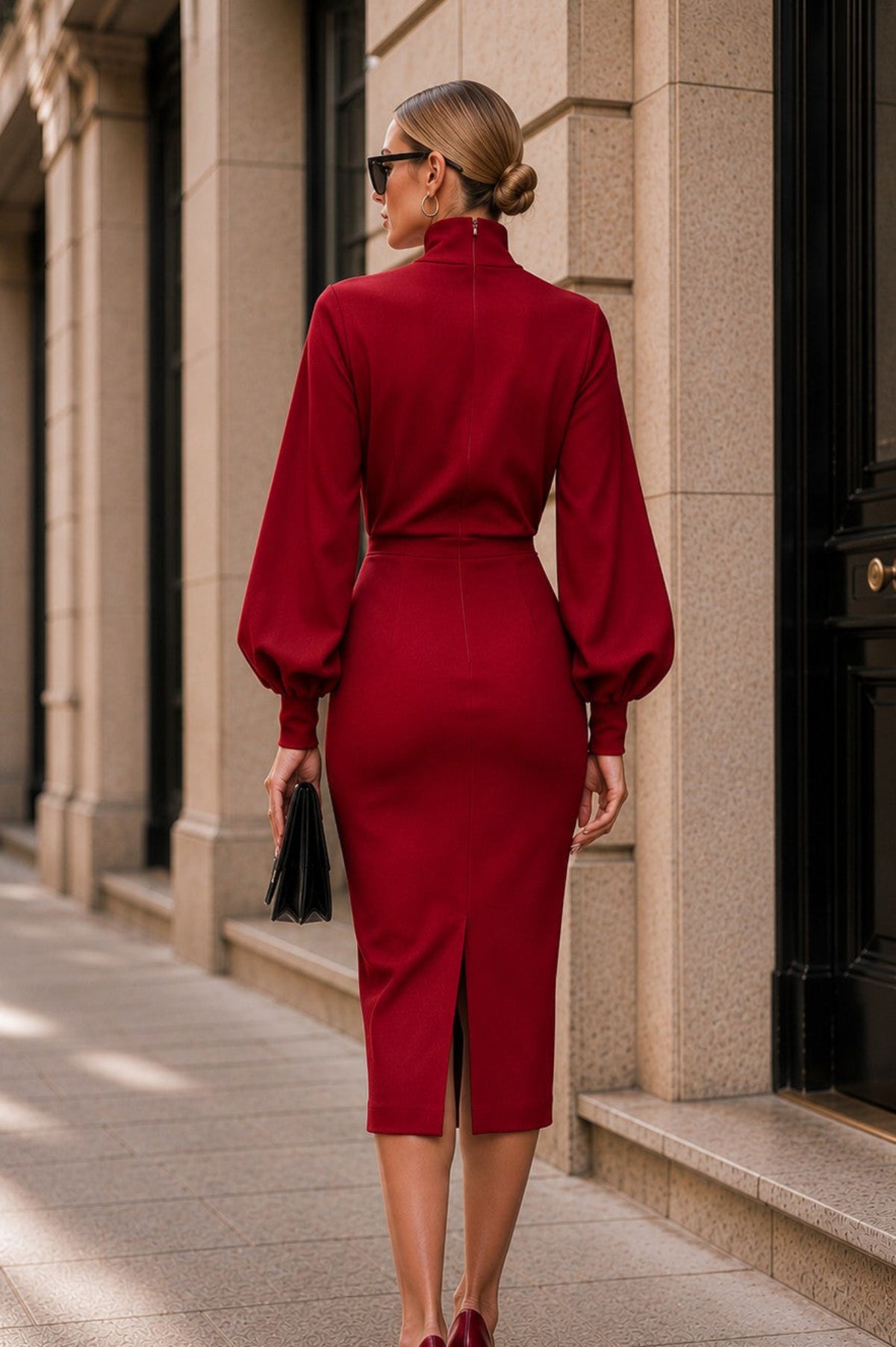 A woman viewed from behind wearing a deep red mock-neck midi dress with balloon sleeves, walking on a city sidewalk next to a stone building.
