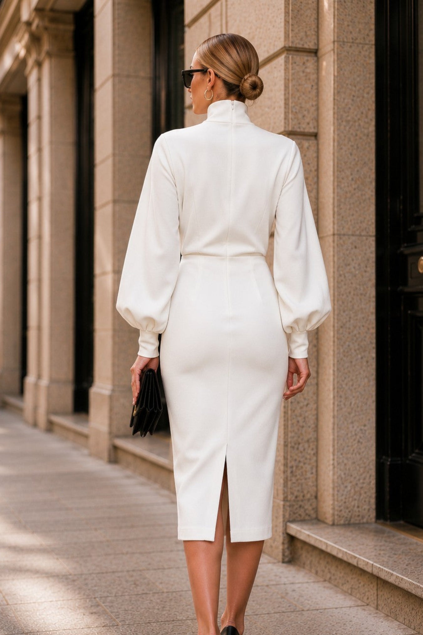 A woman photographed from behind wearing a white high-neck midi dress with balloon sleeves and a front slit, walking on a stone sidewalk beside a building with dark doors.