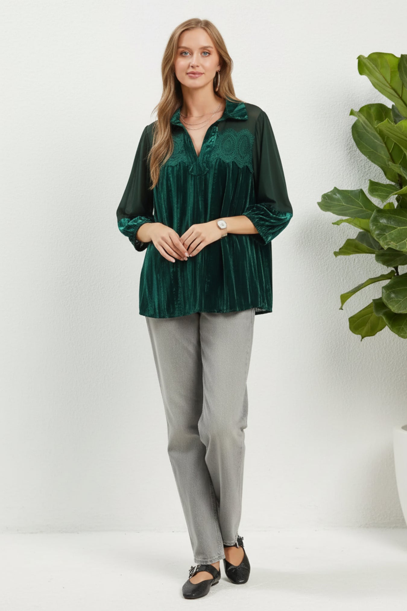 Woman wearing a green velvet blouse and gray pants against a white background next to a plant