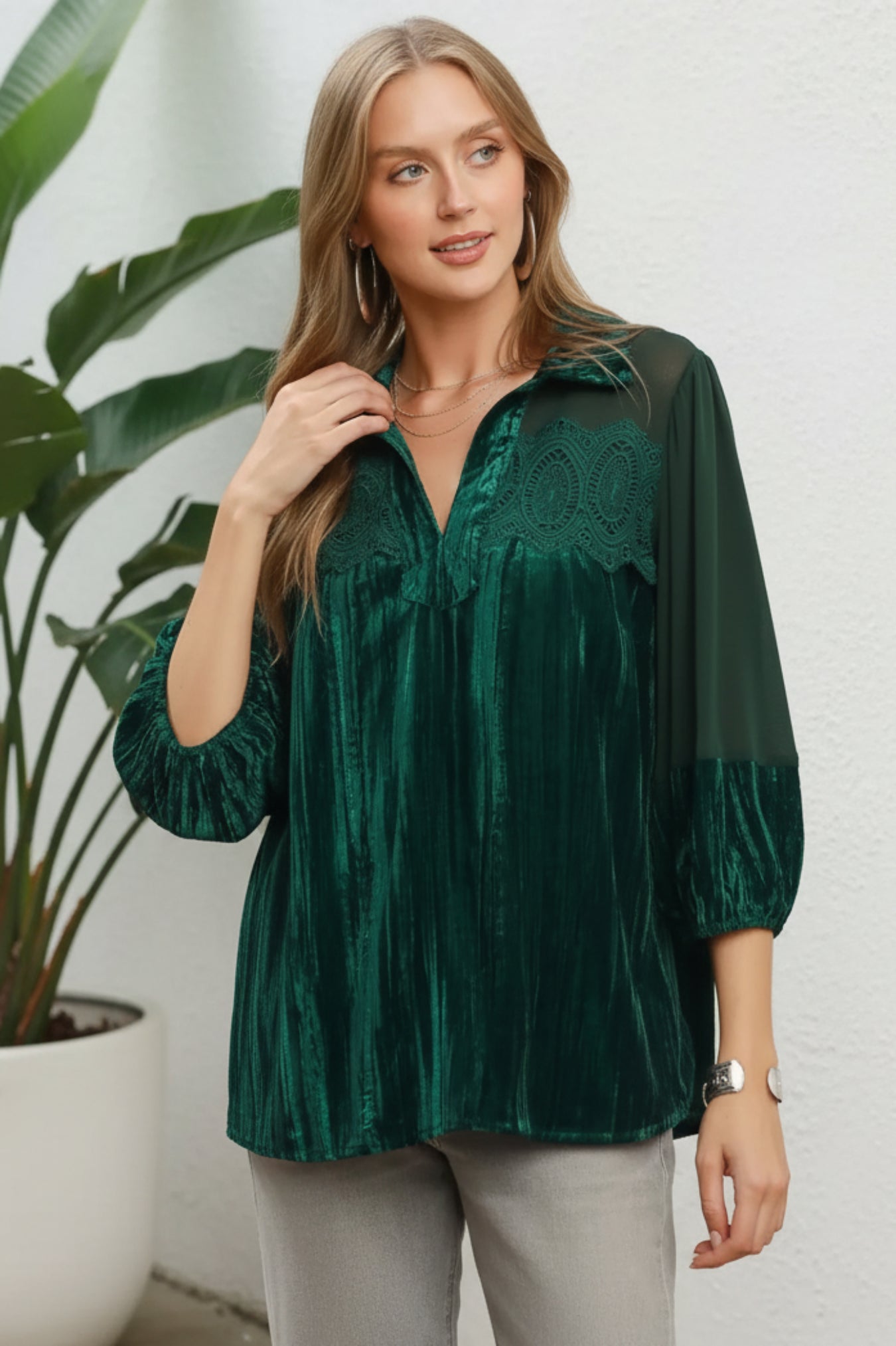 A woman wearing a dark emerald green velvet blouse with sheer mesh sleeves and lace detailing on the chest, posed against a white background and a plant.