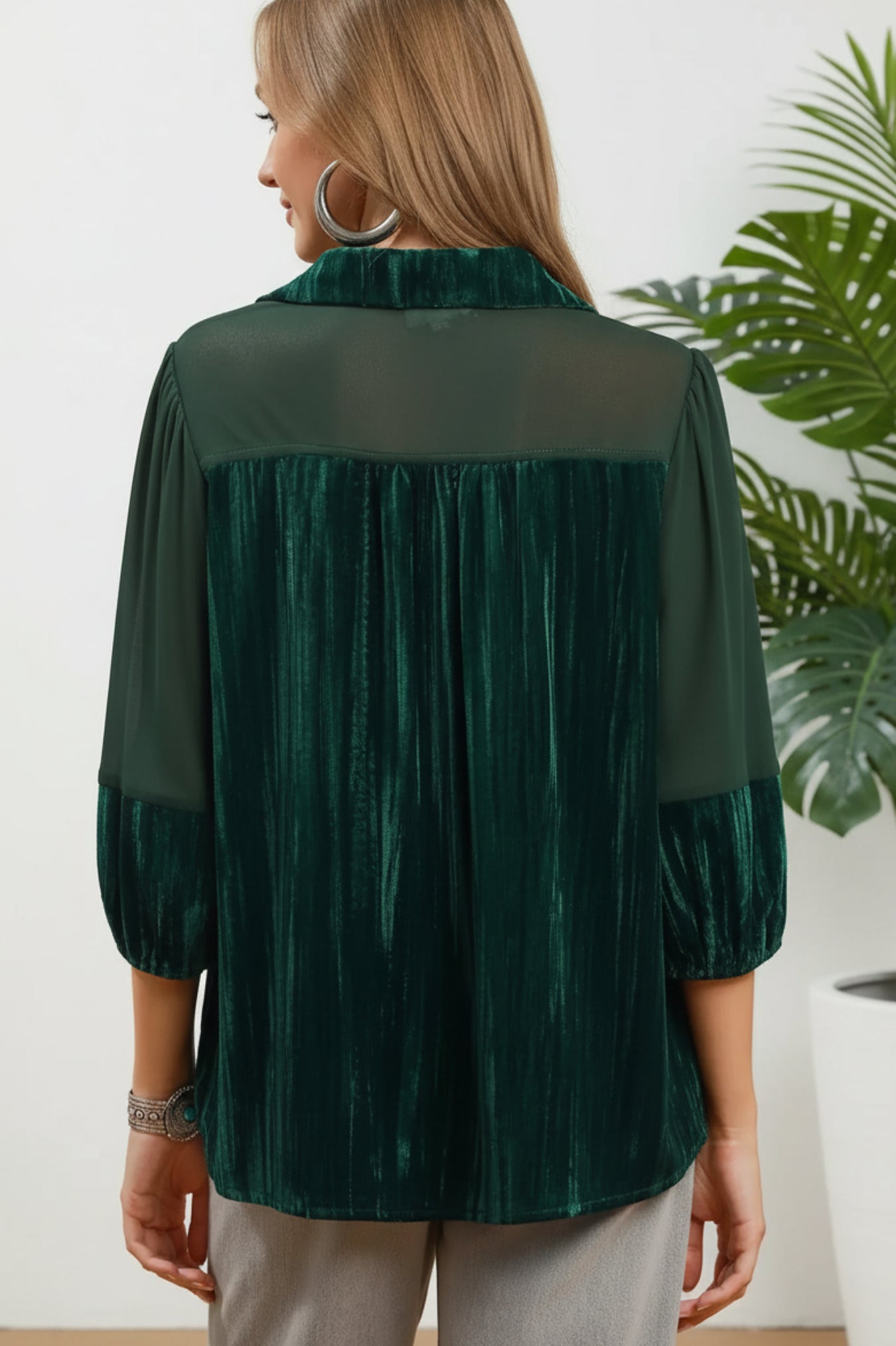 A woman shown from the back wearing a dark green sheer mesh and pleated velvet blouse with three-quarter sleeves, paired with light gray pants, against a white background with a potted plant