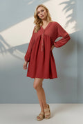A woman modeling a rust-red long-sleeve mini dress with a gathered waist and button placket, paired with beige platform sandals, standing against a light gray wall with soft palm leaf shadows.