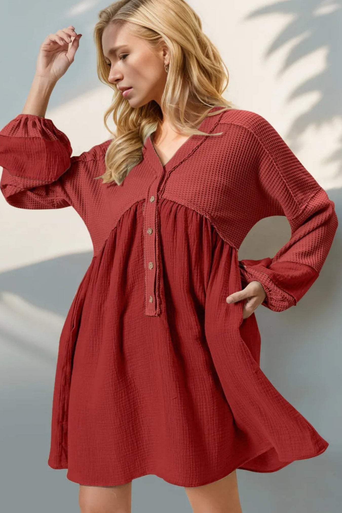 A woman wearing a textured, rust-red long-sleeve babydoll dress with a buttoned yoke and gathered waist, posed against a light gray wall with soft leaf shadows.