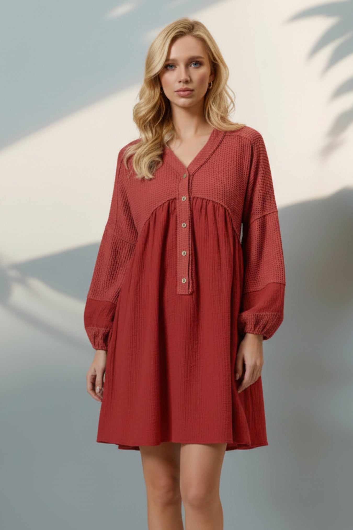 A woman modeling a rust-red crinkle-textured mini dress with long balloon sleeves, a V-neckline, and a gathered waist, paired with beige platform sandals against a gray wall with palm leaf shadows.