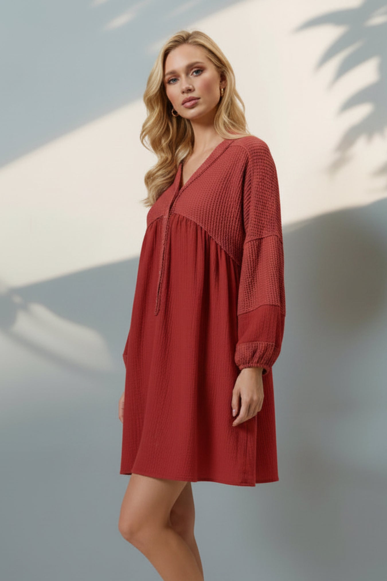 A woman modeling a rust-red long-sleeve mini dress with a gathered waist and button placket, paired with beige platform sandals, standing against a light gray wall with soft palm leaf shadows.